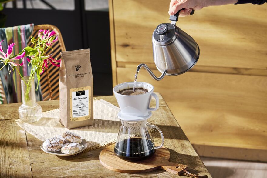Hand gießt Filterkaffee, daneben Bohnen und Gebäck