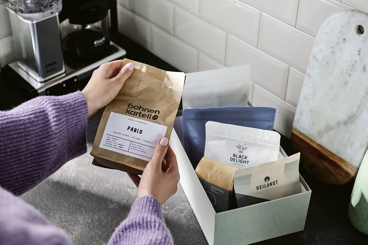 Hände halten Kaffee, Box mit weiteren Kaffeepackungen