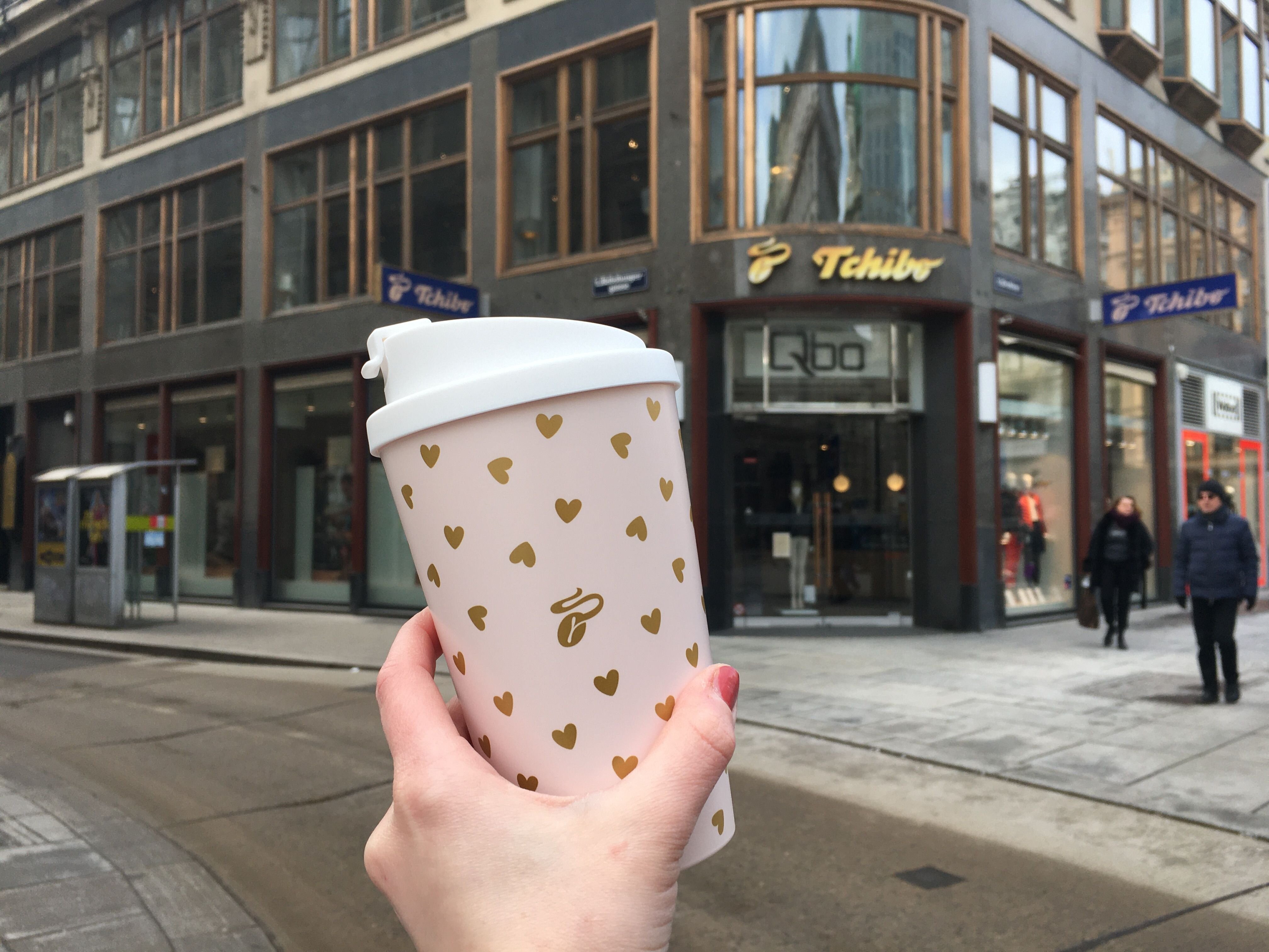 Hand hält Becher mit Herzchen vor Tchibo-Filiale