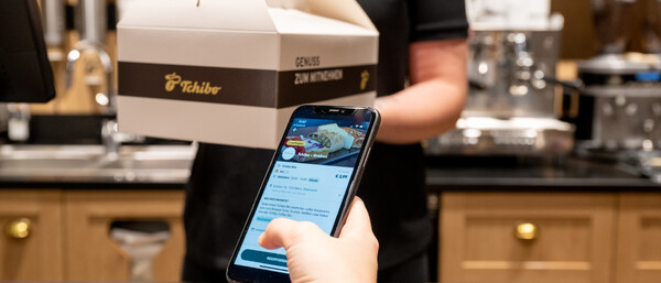 Kundin zeigt Smartphone, Barista reicht Tchibo-Box
