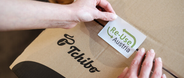 Hände kleben Re-Use Austria Sticker auf Tchibo Karton