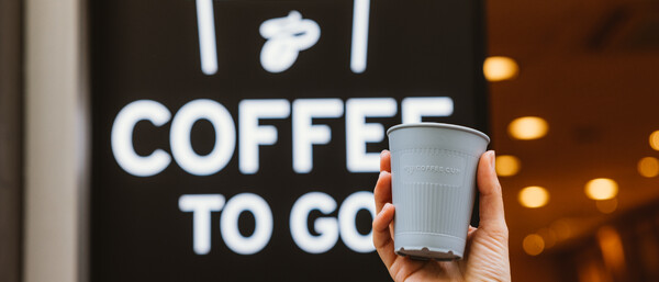 Hand hält grauen Mehrwegbecher vor „COFFEE TO GO“-Schild