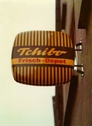 Link: Großansicht des Bildes - Bildinhalt: Außenaufnahme eines leuchtenden, gelb-braun gestreiften Schilds mit abgerundeten Kanten, an dem in braunen Lettern „Tchibo“ und darunter in roter Schrift „Frisch-Depot“ steht.