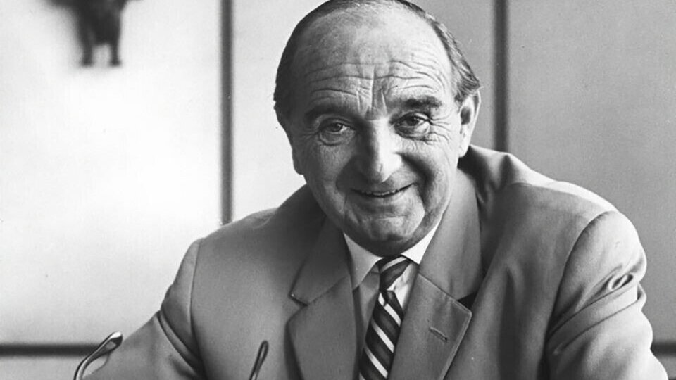 Mann im Anzug mit Krawatte sitzt am Schreibtisch, hält seine Brille in der Hand, im Hintergrund eine afrikanische Maske.