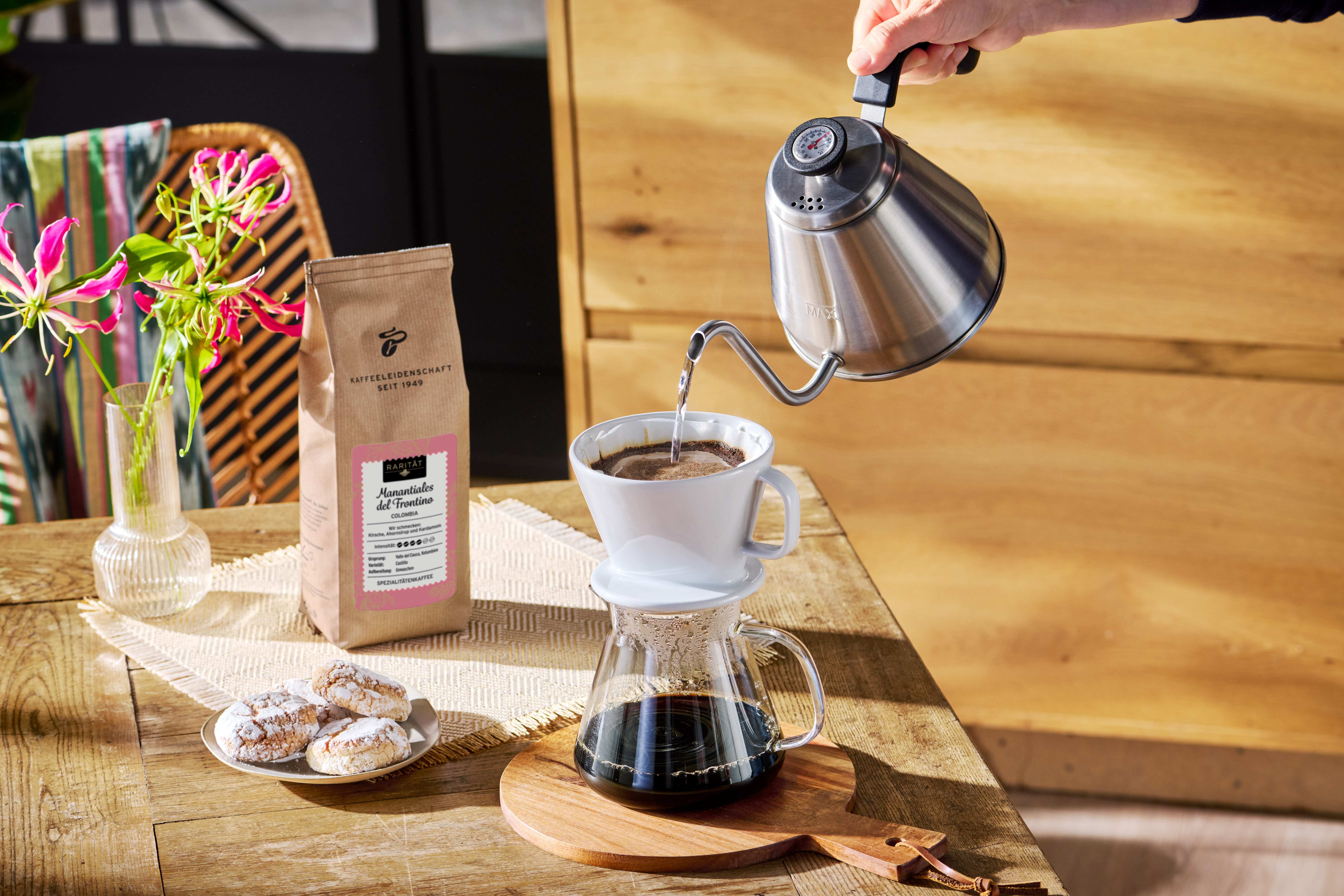 Hand gießt Filterkaffee neben Bohnenpackung und Gebäck