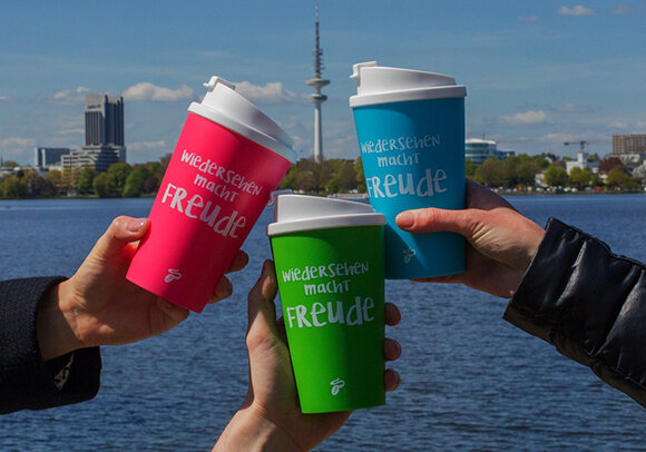 Drei bunte Becher mit Schriftzug vor Stadtsee