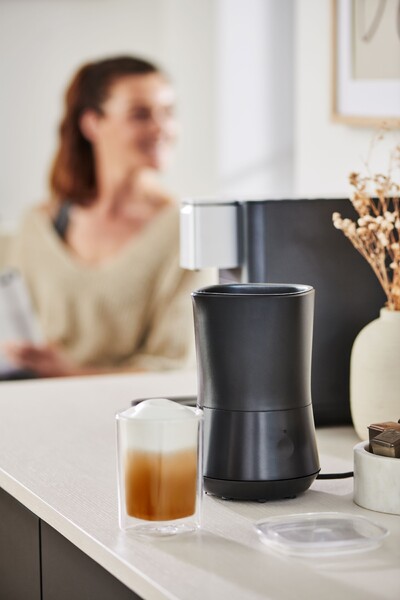 Milchaufschäumer mit Kaffee neben unscharfer Frau