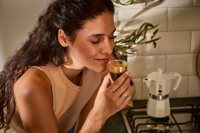 Frau hält Espresso neben Herd mit Espressokocher