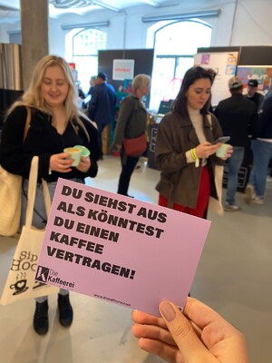 Link: Großansicht des Bildes - Bildinhalt: Hand hält lila Karte mit Kaffeewerbung in Messehalle