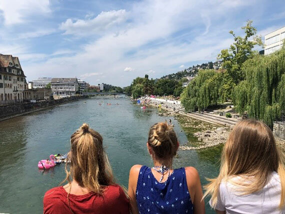 Drei Mädchen blicken auf einen Fluss mit Badenden