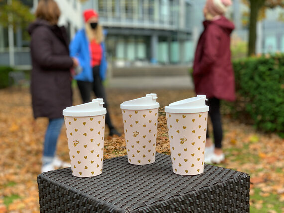 Drei Kaffeebecher, dahinter drei Personen draußen