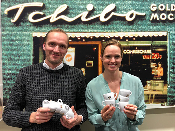 Zwei Personen halten Tchibo-Kaffeetassen vor Ladenfassade