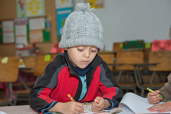 Kind mit Wollmütze malt mit Buntstiften im Klassenraum