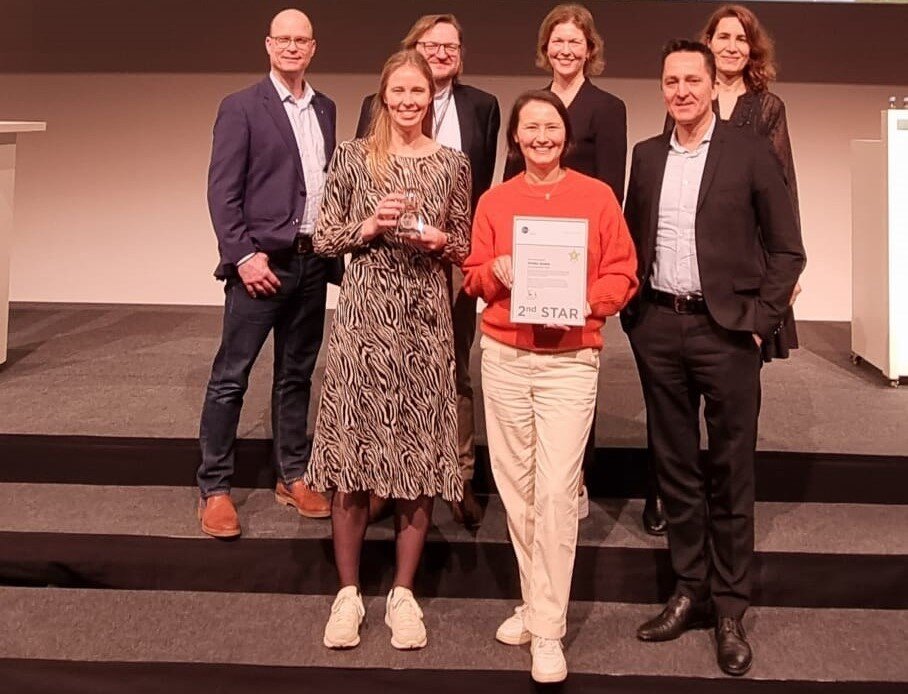 Gruppe auf Bühne, Frau hält Urkunde „2nd STAR“