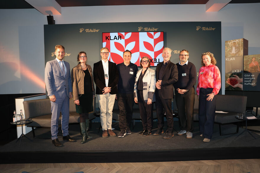 Acht Personen auf Podium vor Tchibo‑Wand