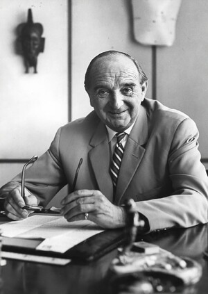 Link: Large view of image - Imagecontent: Man in a suit and tie sitting at a desk, holding his glasses in his hand, an African mask in the background.